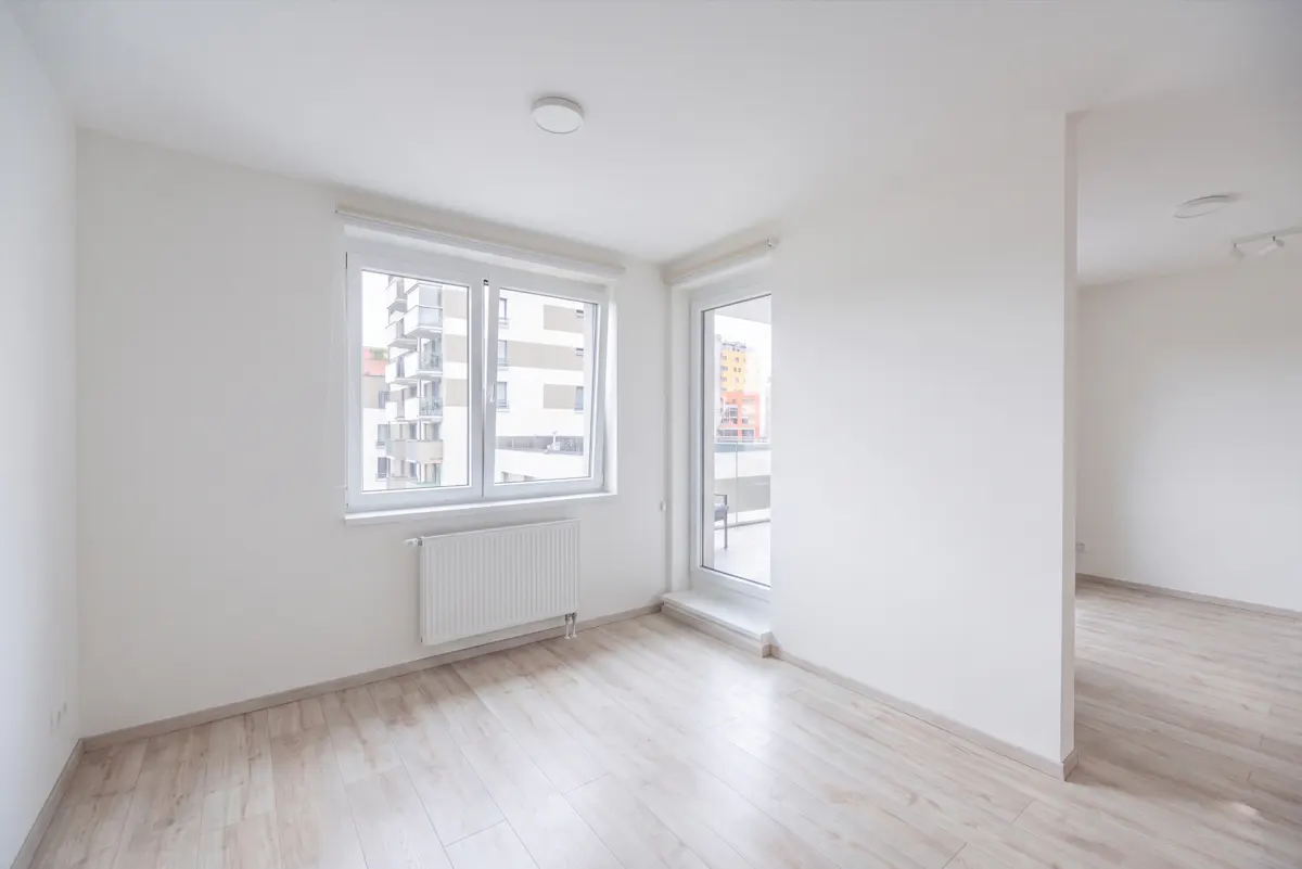 Empty room with white walls, light wood floors, a window with a radiator, and a glass door leading to a balcony.