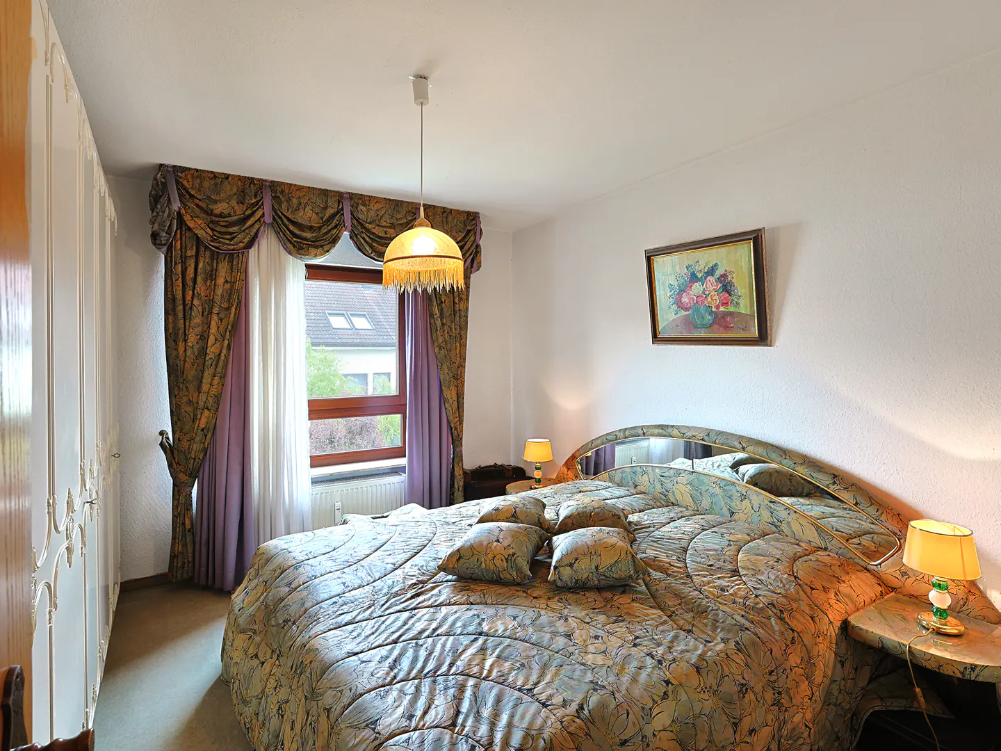 Bedroom with a large round bed, floral bedding, and matching pillows. A window with floral curtains and a painting of flowers on the wall.