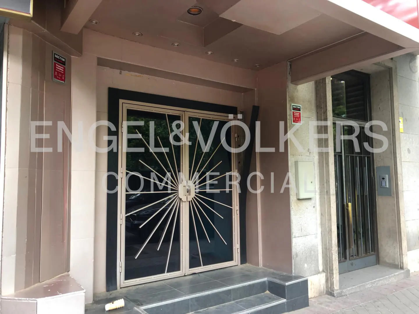 Exterior view of a commercial building with black framed doors and a sunburst design. The Engel & Volkers logo is superimposed on the image.