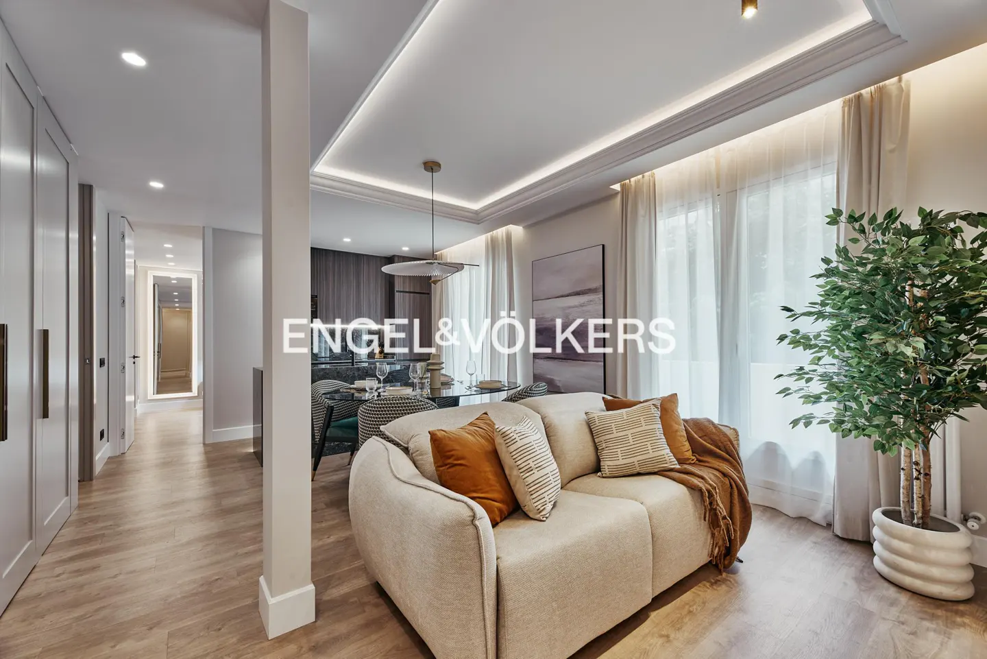 Living room with beige sofa, brown pillows, and a plant near the window. Dining table and chairs are in the background.
