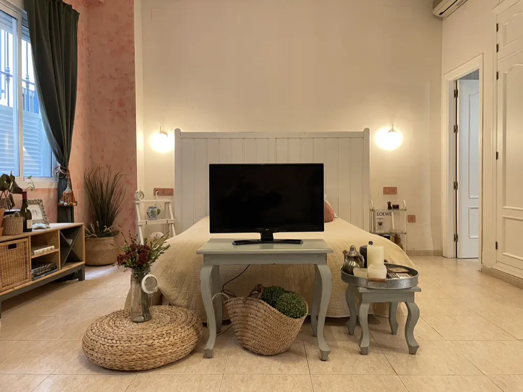 A bedroom with a white headboard, a TV on a small table, a woven floor pouf, and decorative accents on a tiled floor.