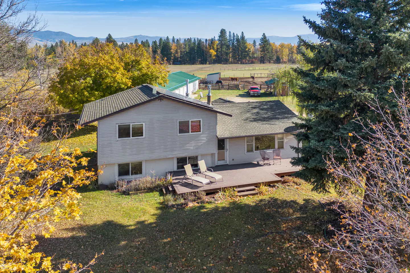 Two-story gray house with a deck and lawn chairs, set against a backdrop of trees, fields, and mountains.