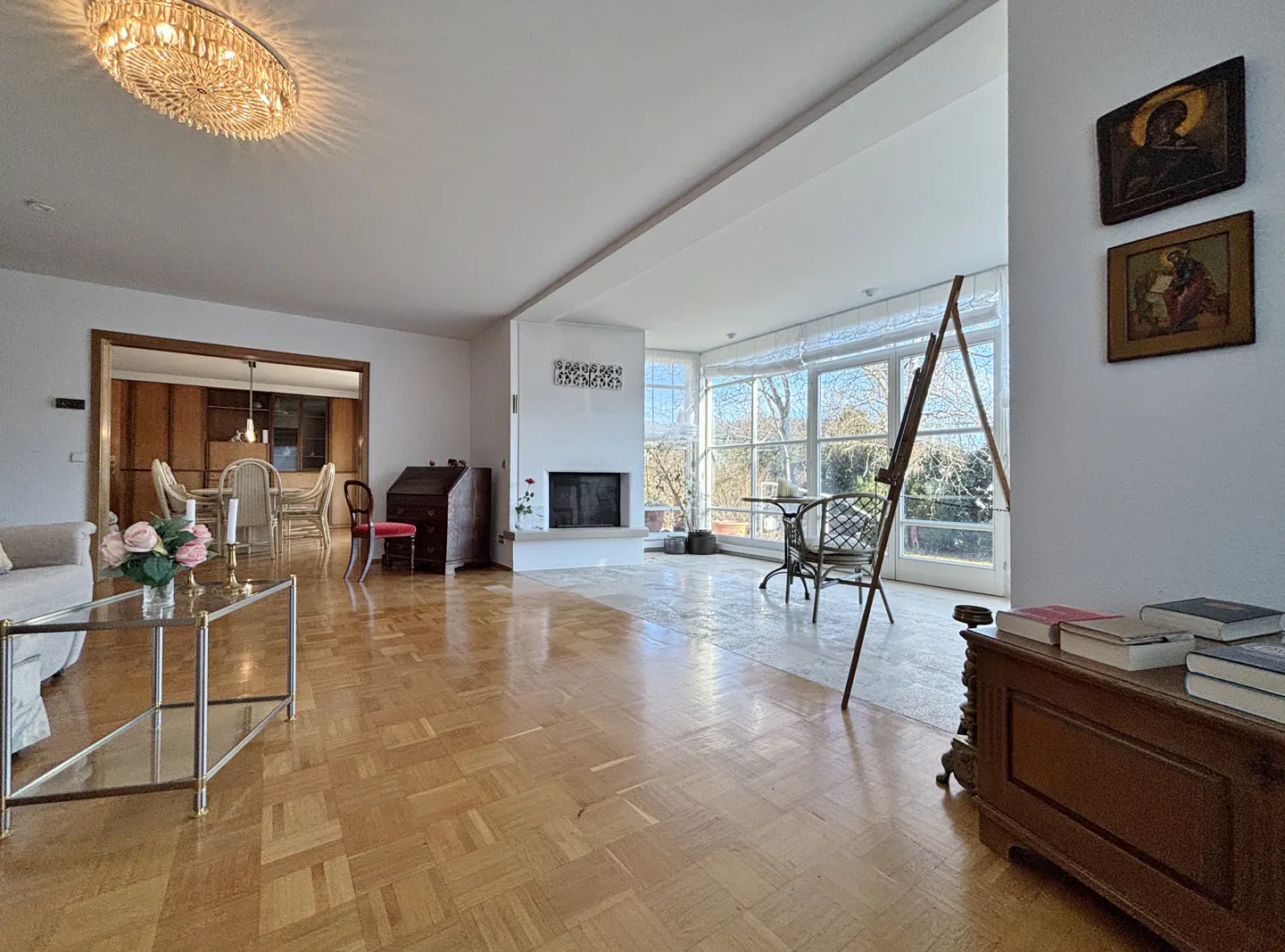 Bright living room with parquet floors, a fireplace, and large windows. An easel stands near a chair, and religious art hangs on the wall.