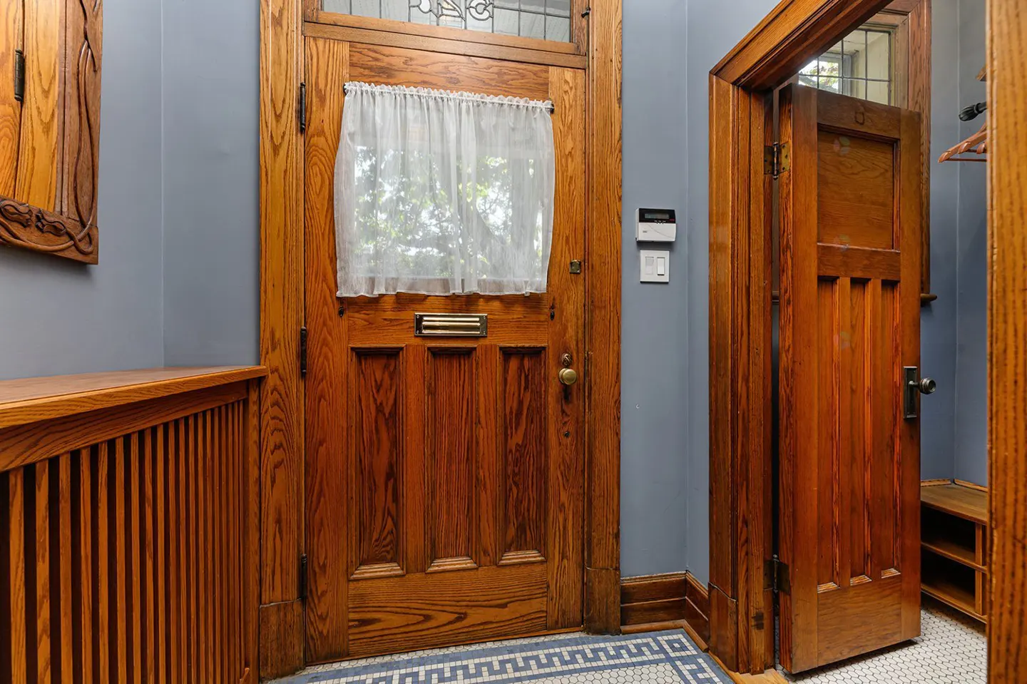 Interior shot of a foyer with a wooden front door with a white curtain and a mail slot. An open wooden door is to the right. Blue walls and tiled floor.