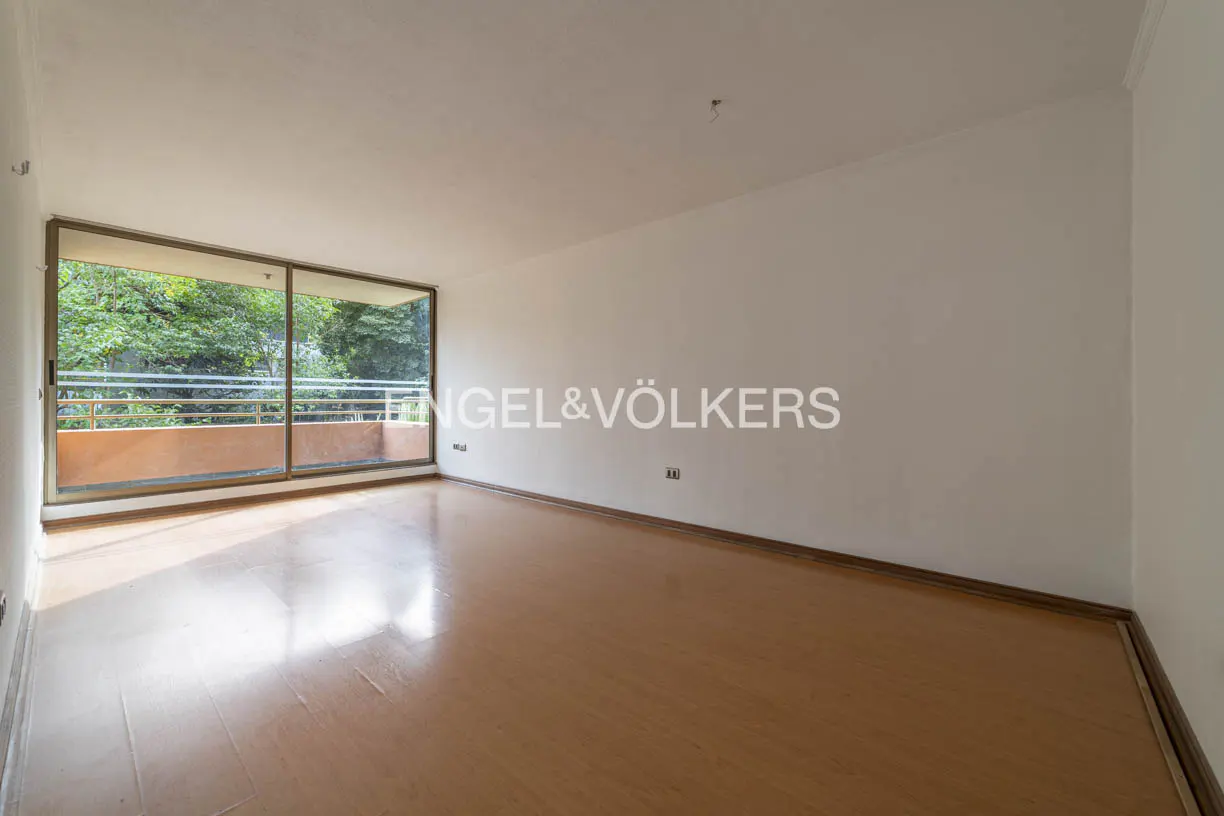 An empty room with wood floors and white walls. A large sliding glass door leads to a balcony with trees visible. Engel & Völkers logo on the wall.