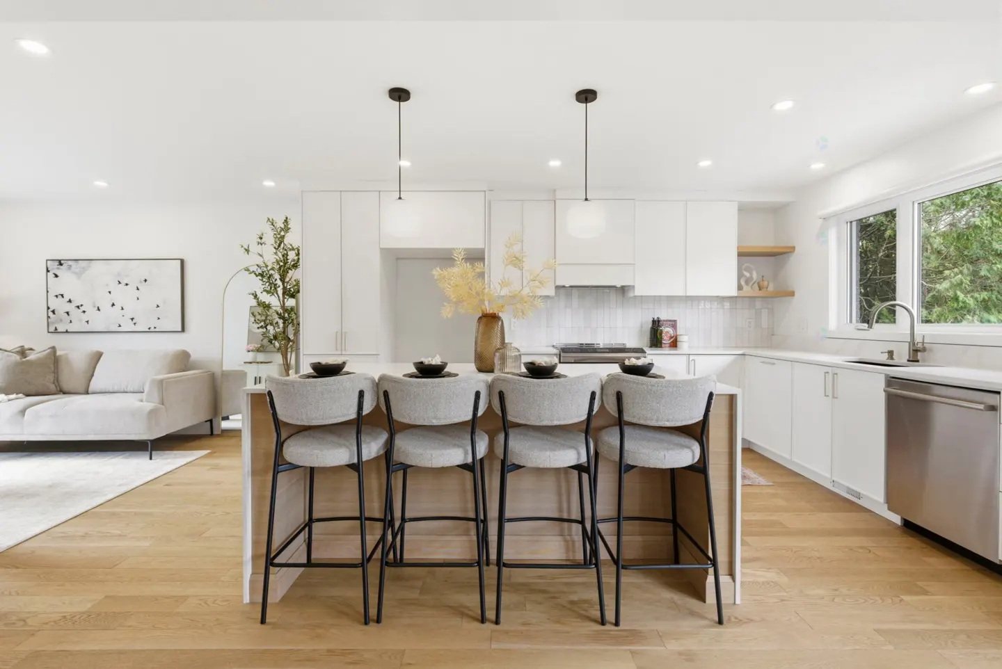 Bright, modern kitchen with white cabinets, stainless steel appliances, and a wood floor. Four gray chairs line the island. Living room in background.