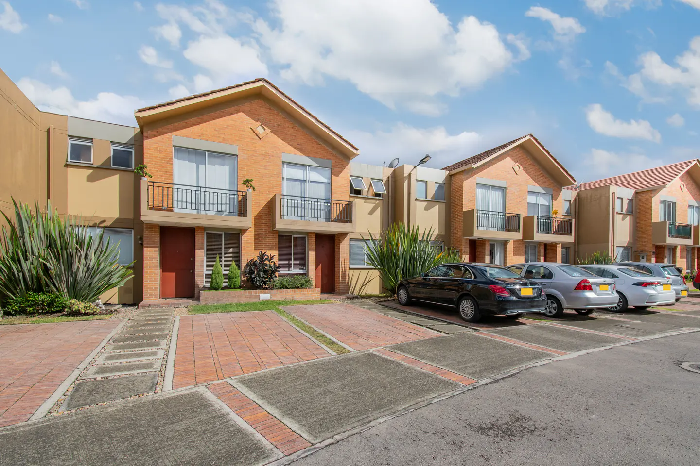 Row of two-story townhouses with brick and tan exteriors, small balconies, and parked cars under a blue, cloudy sky.