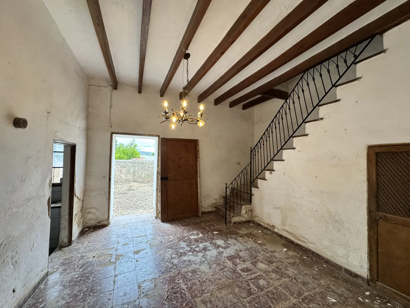 Interior of a rustic room with white walls, exposed wood beams, a chandelier, and a staircase with black railings.