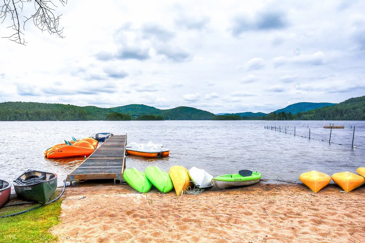 Lake view with a dock, boats, and kayaks on a sandy beach under a cloudy sky. Mountains are visible in the background.