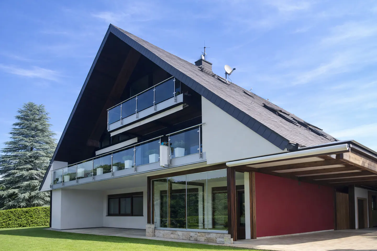 Modern two-story house with a dark gray roof, white walls, glass balconies, and a red accent wall under a wooden carport.