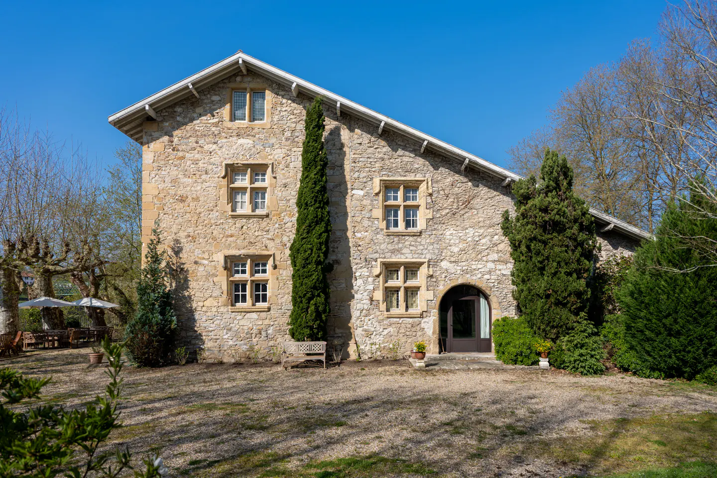 Exterior view of a two-story stone house with a gray roof and arched doorway, surrounded by trees and a lawn.