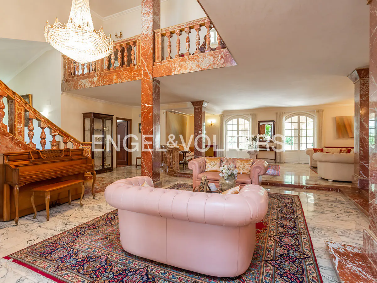 Opulent living room with pink sofas, marble floors, and a grand staircase with a piano. A large chandelier hangs from the high ceiling.