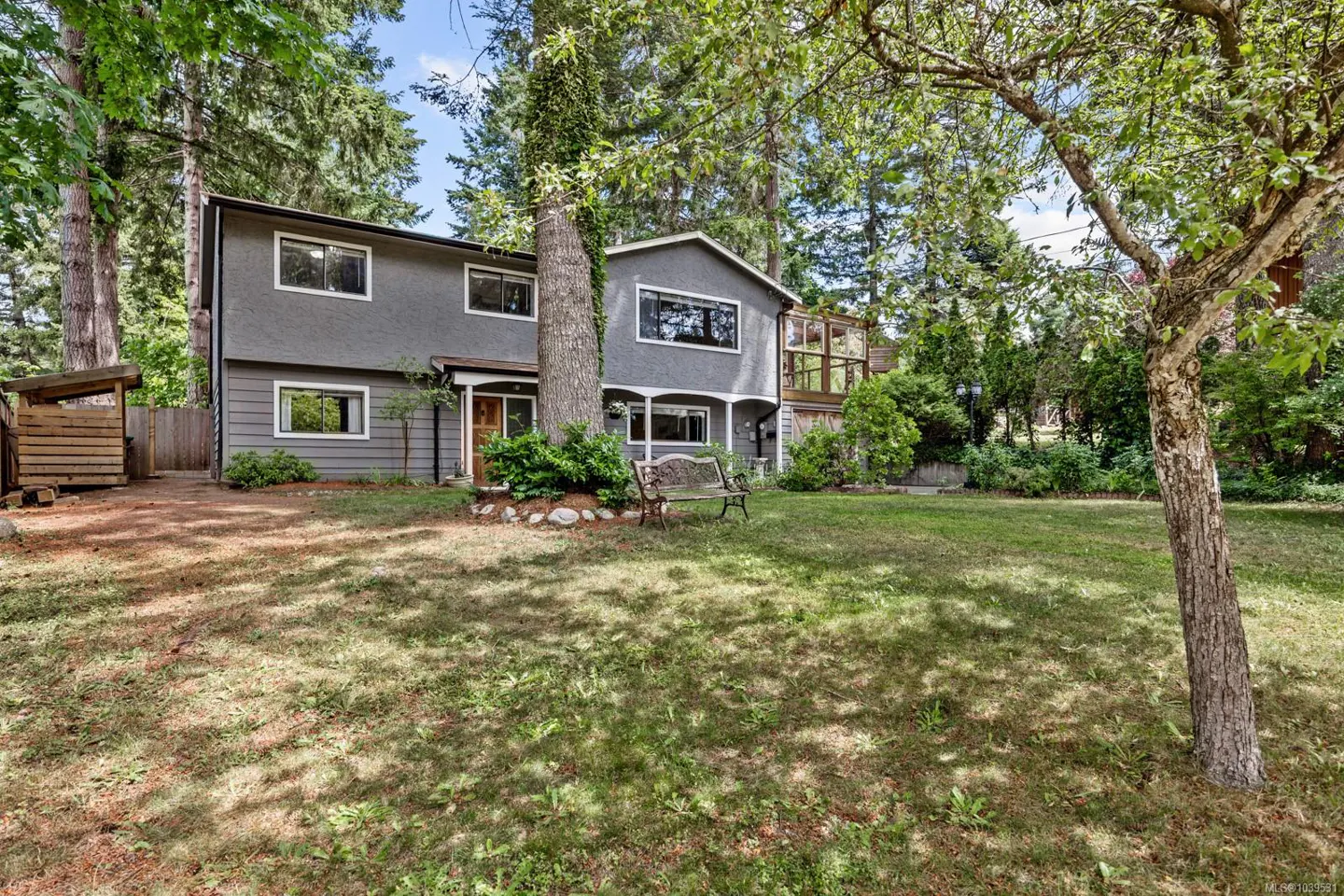 Two-story grey house with a wooden bench in a large, tree-filled yard with a small shed on the left.