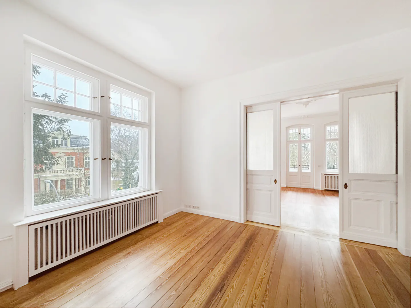 Bright, empty room with wood floors, white walls, and large window. Sliding doors open to another room with windows.