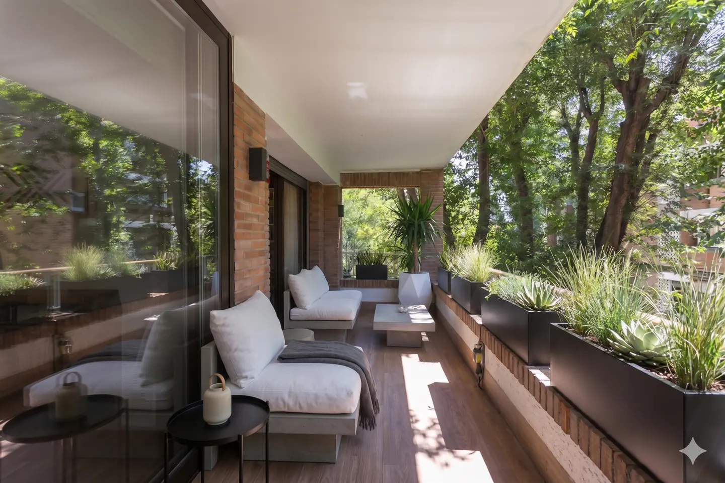 Modern balcony with white cushioned seating, a small coffee table, and black planters filled with greenery and trees.