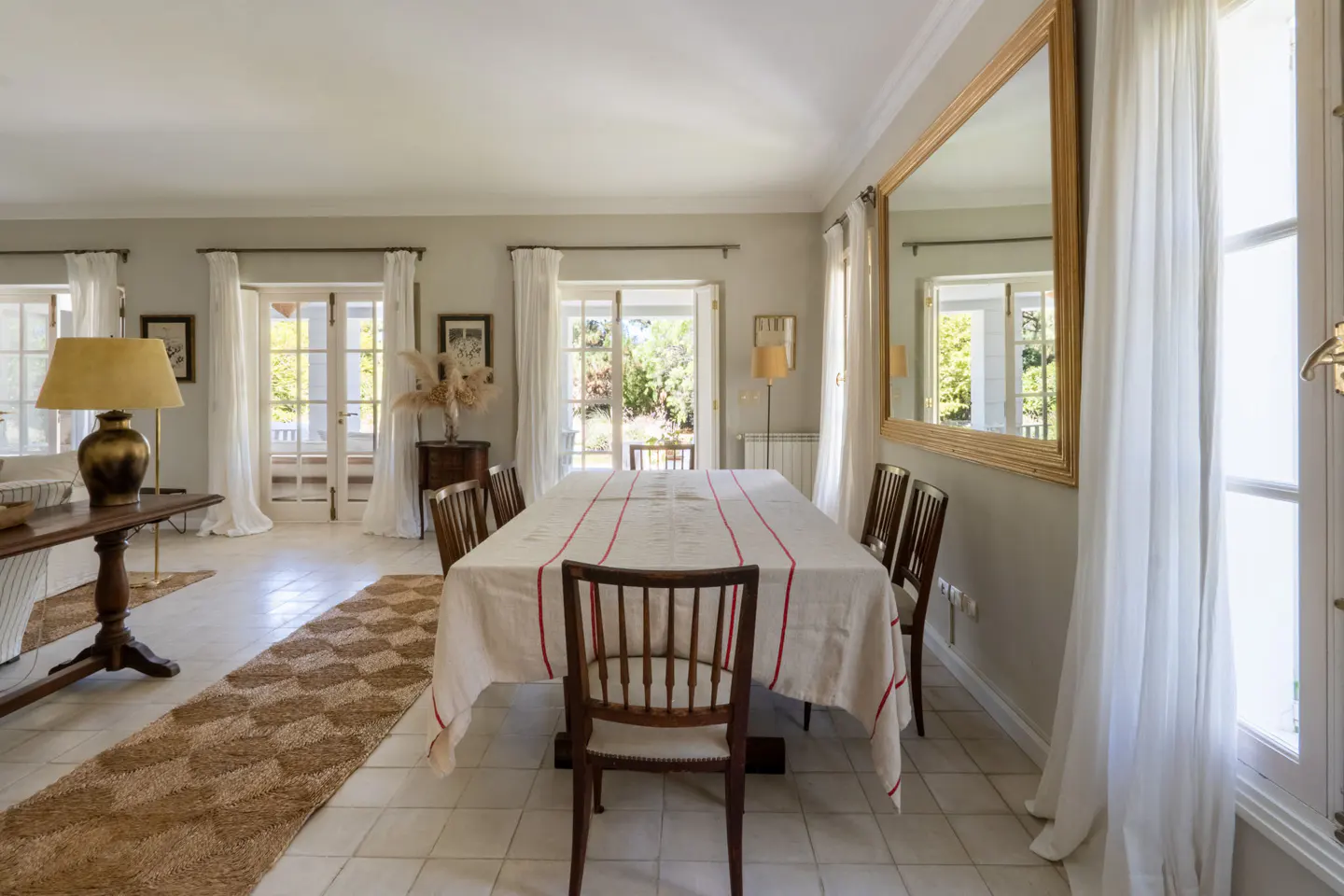 Bright dining room with a long table covered in a white tablecloth with red stripes, surrounded by wooden chairs. Large windows with white curtains.