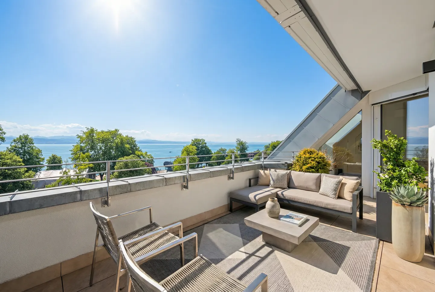 A sunny balcony with seating overlooks a blue lake and green trees. Two chairs face a sofa, table, and potted plants.