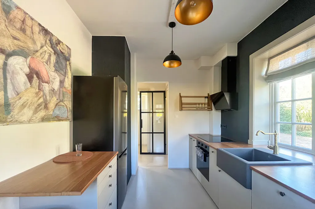 A modern kitchen with white cabinets, wood countertops, and black accents. A large painting hangs on the wall.