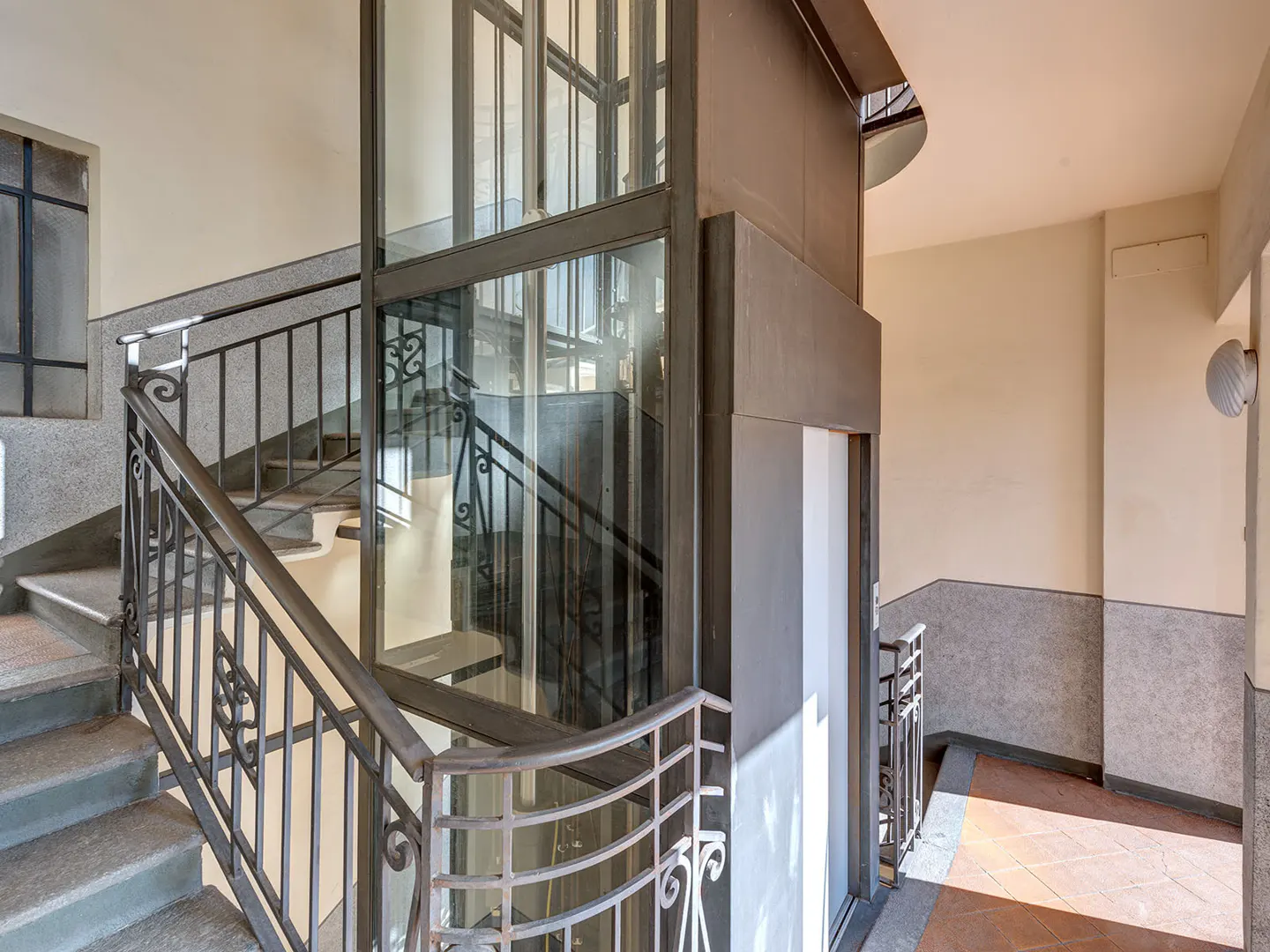 Interior view of a building with stairs and a glass elevator. The walls are cream and gray, and the floor is tiled.