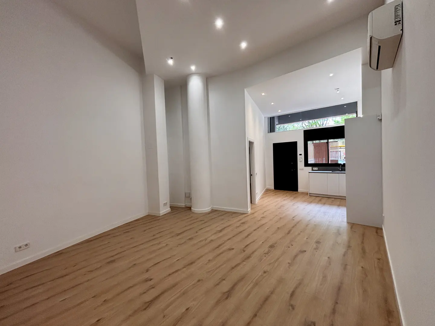 Bright, empty room with wood floors, white walls, and recessed lighting. A black door and white kitchen cabinets are visible in the background.