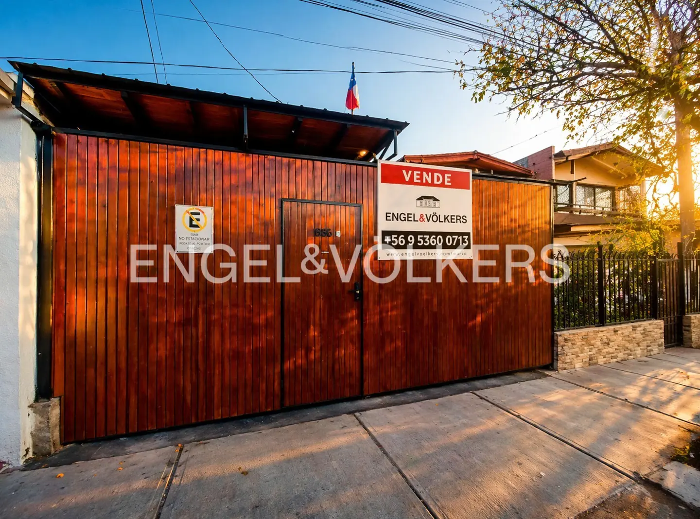 Exterior shot of a house with a large wooden gate and an Engel & Völkers "For Sale" sign. A Chilean flag is on top of the gate.