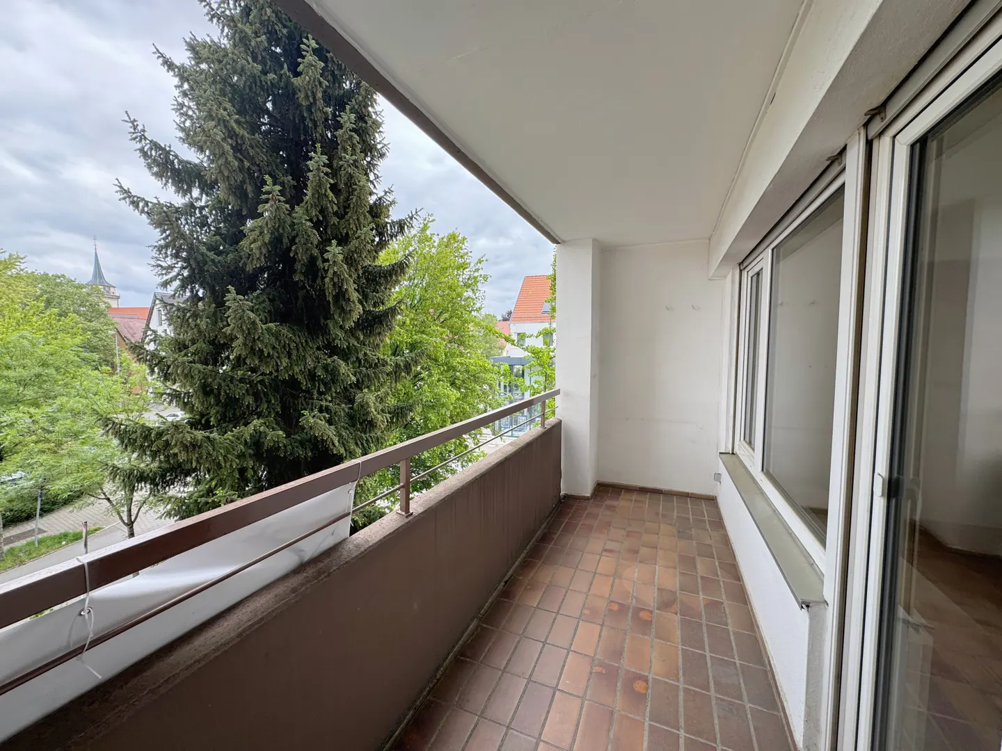 A balcony with brown tiled floor and railing overlooks green trees and a church steeple in the distance. White walls and windows frame the view.