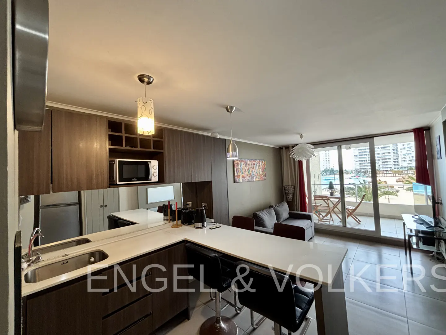 Open-concept apartment interior with a kitchen island, gray sofa, and balcony overlooking a beach.