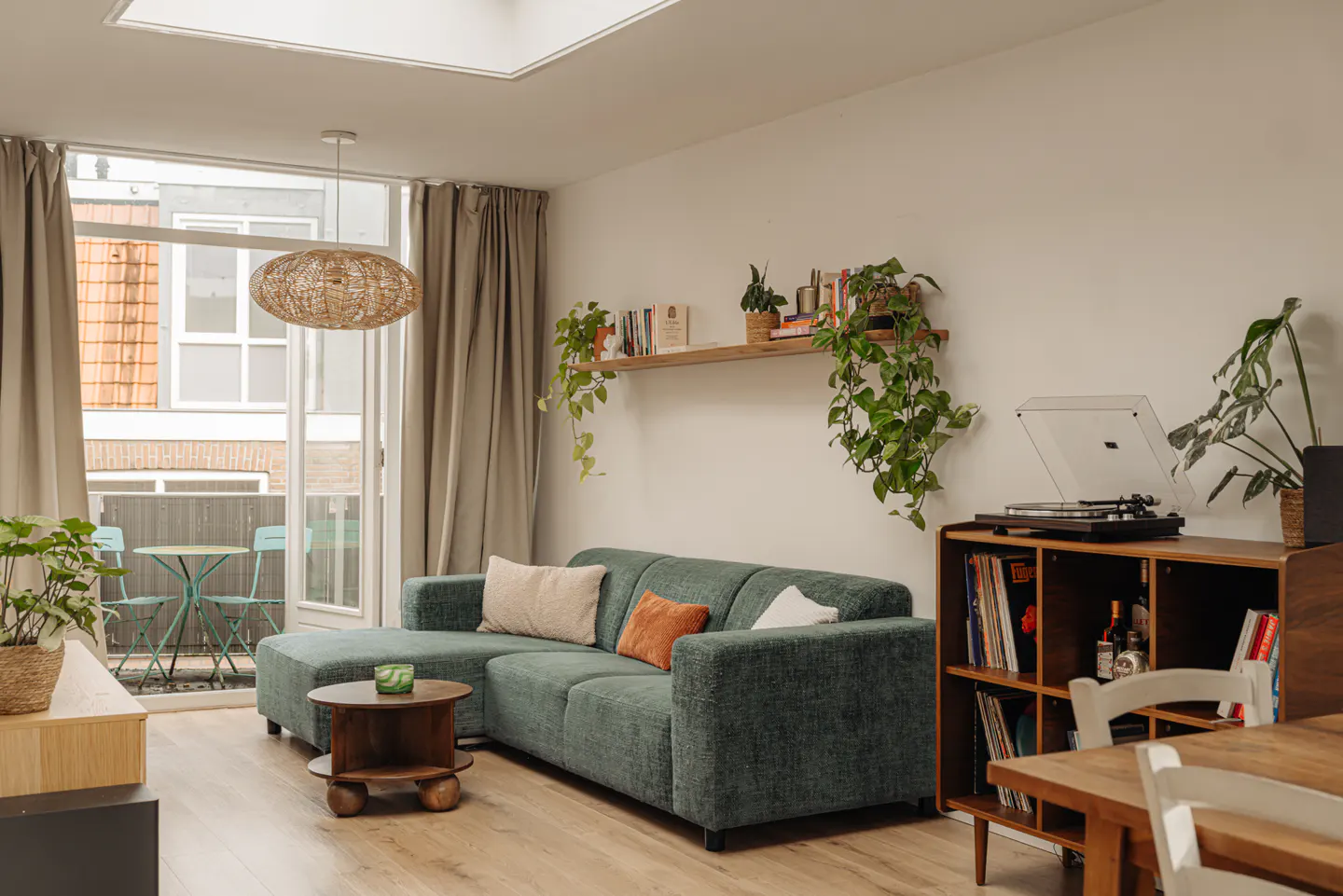 Bright living room with a green sofa, wooden shelf with plants, and a record player on a wooden cabinet. Balcony with table and chairs visible.