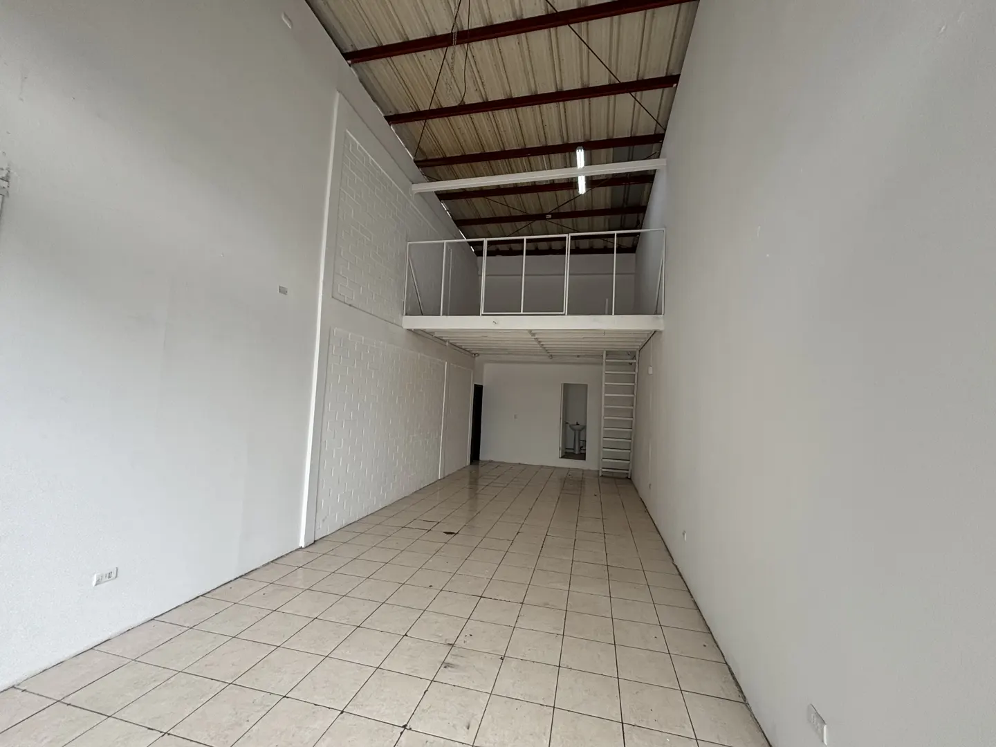 Empty commercial space with white walls, tile floor, and a metal roof. A white metal mezzanine is accessible by a ladder.