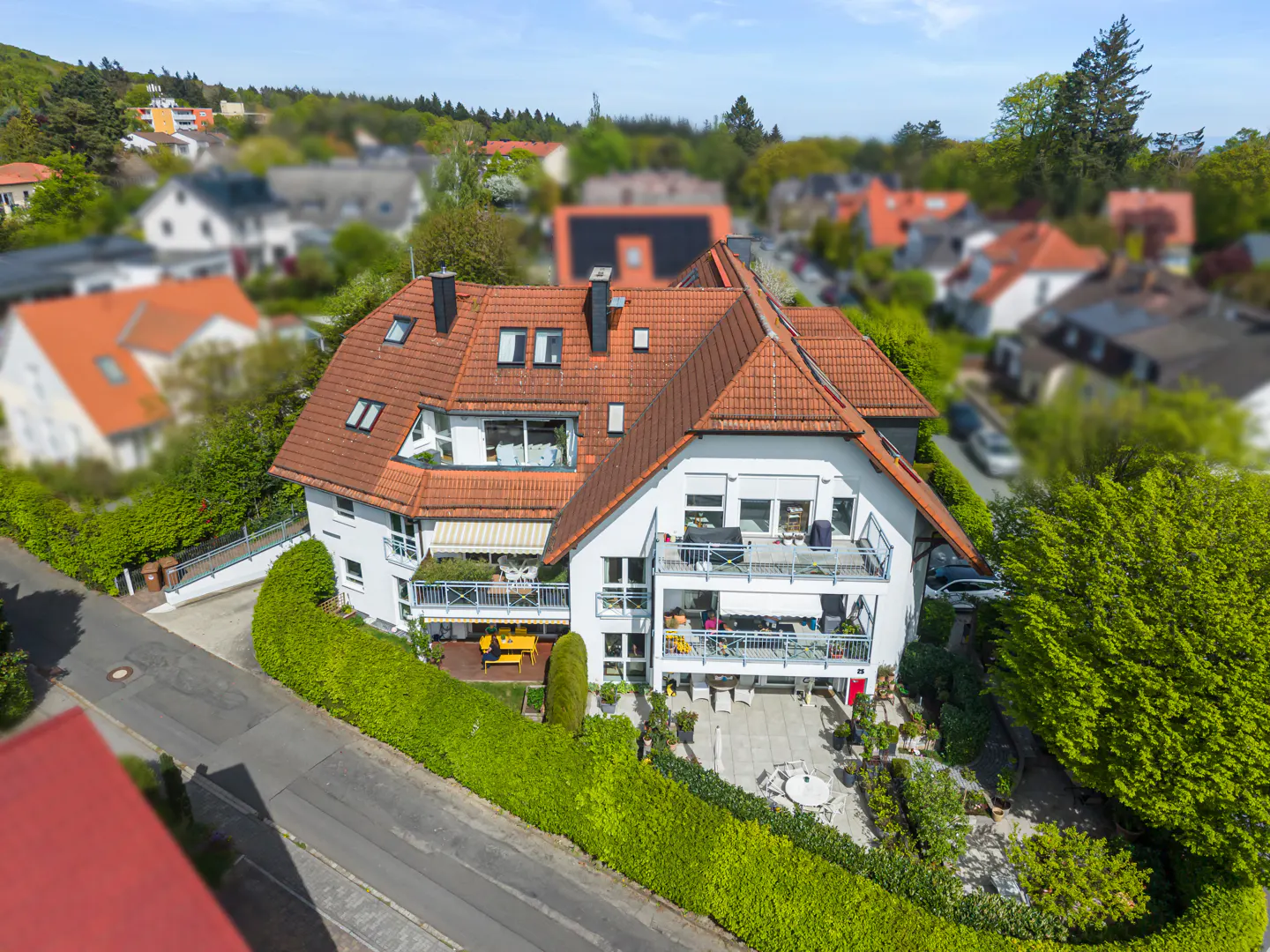 Aerial view of a white house with a red tile roof, multiple balconies, and a green hedge surrounding the property.