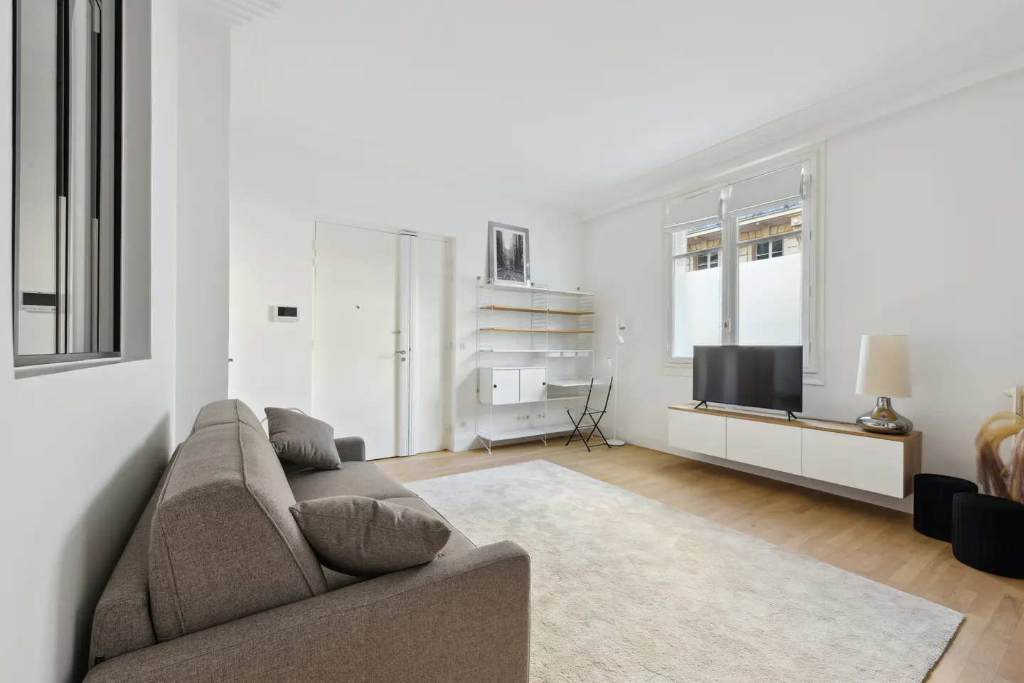 Bright living room with a gray sofa, white walls, and light wood floors. A white shelving unit and TV stand add modern touches.