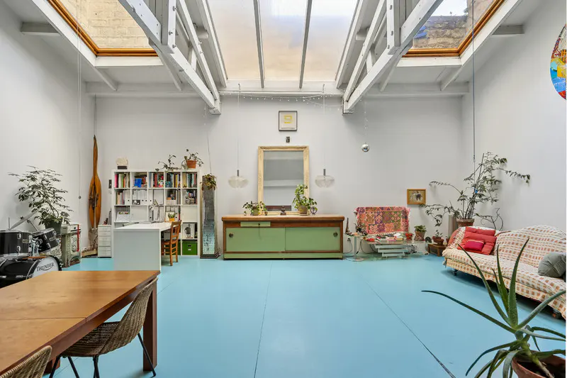 Bright, airy living space with blue floors, white walls, and skylights. Features a wooden table, sofa, desk, and vintage cabinet.