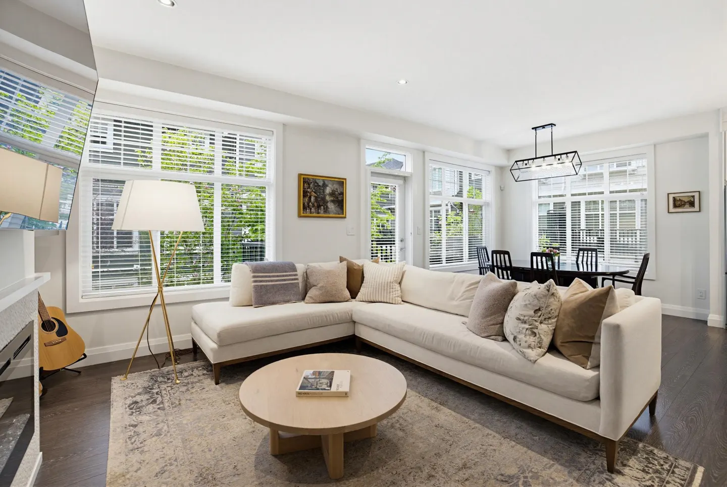 Bright living room with white walls, a large sectional sofa, and a round coffee table on a patterned rug. A dining table is visible in the background.