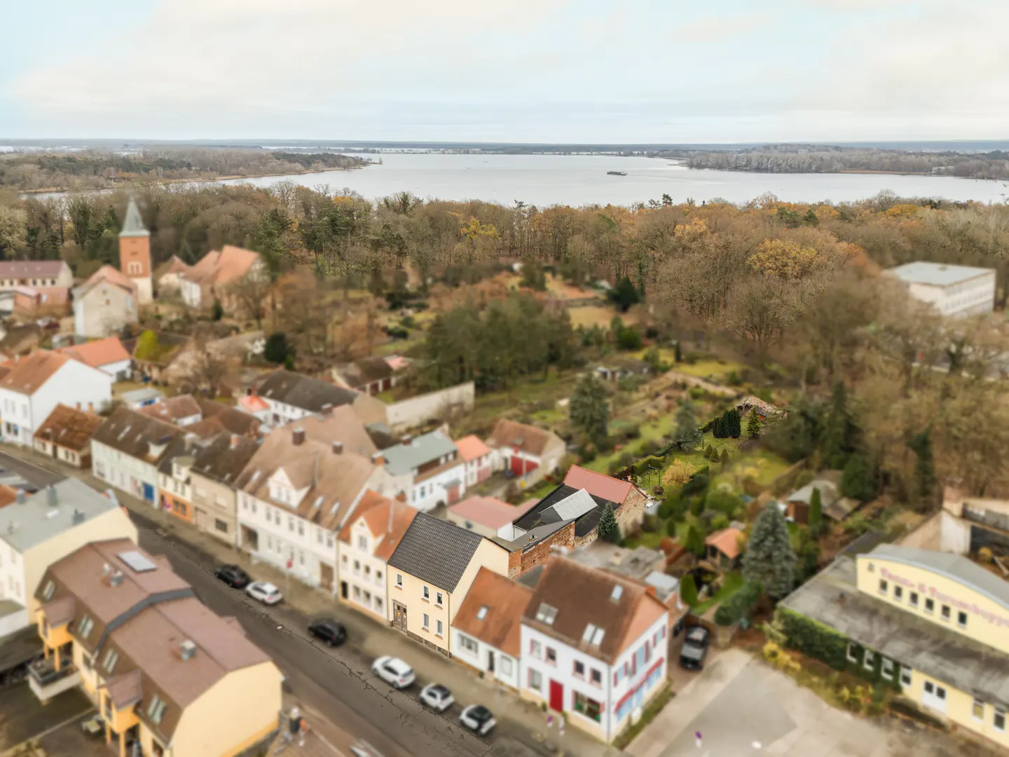 Aerial view of a European town with red-roofed buildings, a church, and a lake in the background. Cars line the street.
