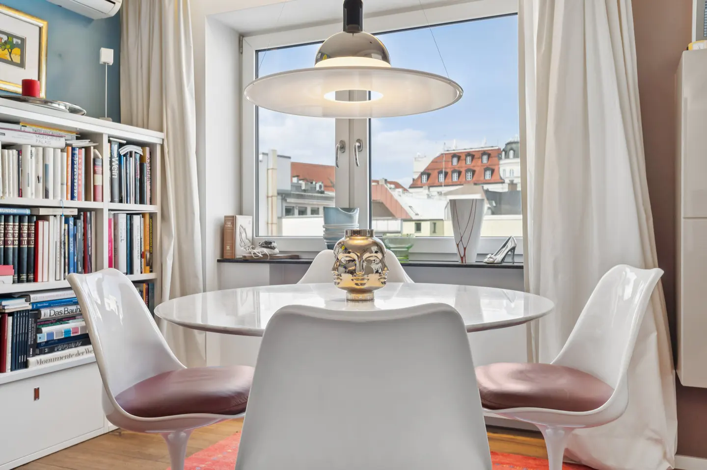 Bright dining area with white tulip chairs around a round table, lit by a modern pendant lamp. A bookcase and city view add to the ambiance.
