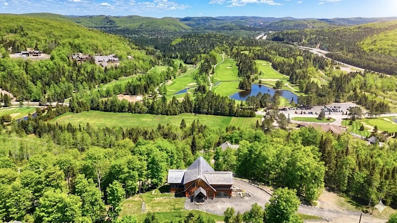 Aerial view of a golf course and a brown building with a gray roof surrounded by green trees and mountains.