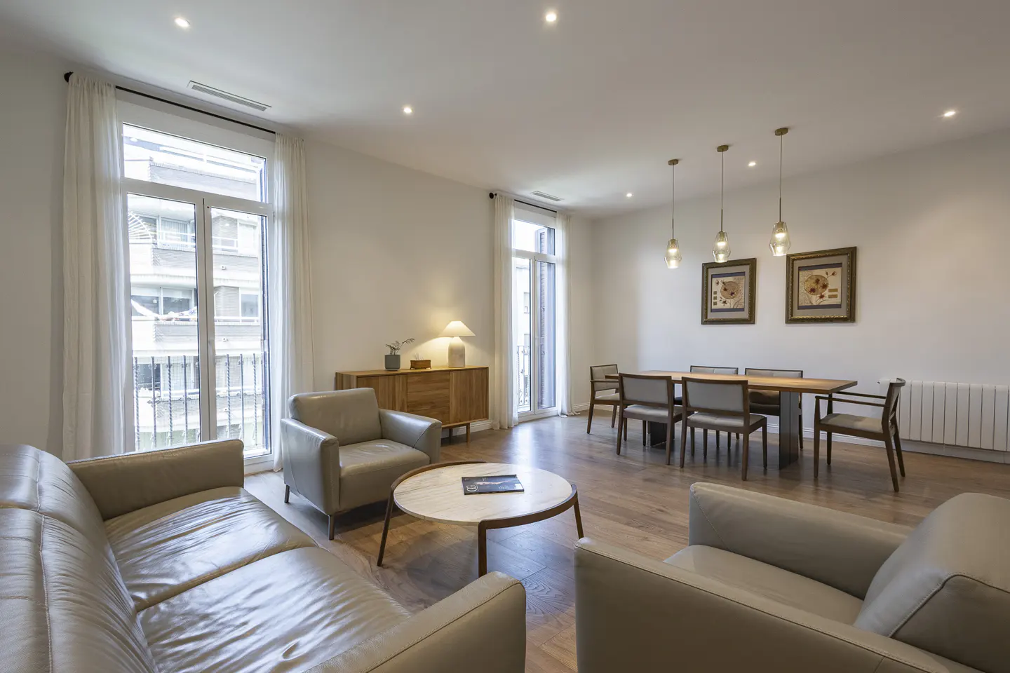 Bright living room with hardwood floors, beige leather sofa and chairs, dining table with six chairs, and large windows with white curtains.