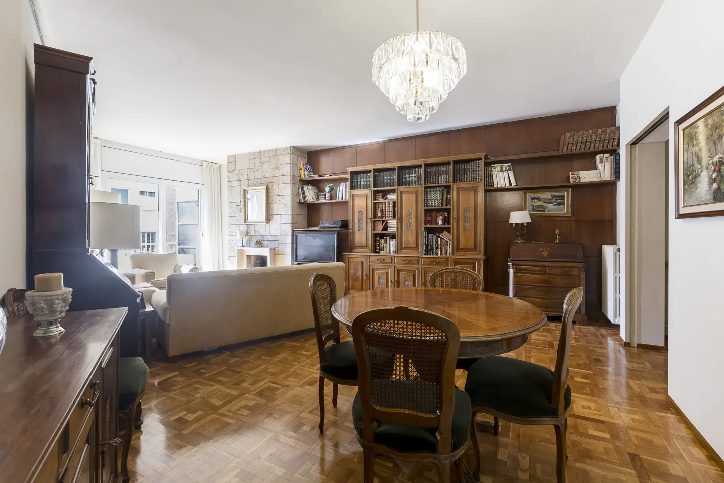 A living and dining area with wood floors, a round table with chairs, and a large wooden bookcase. A crystal chandelier hangs from the ceiling.