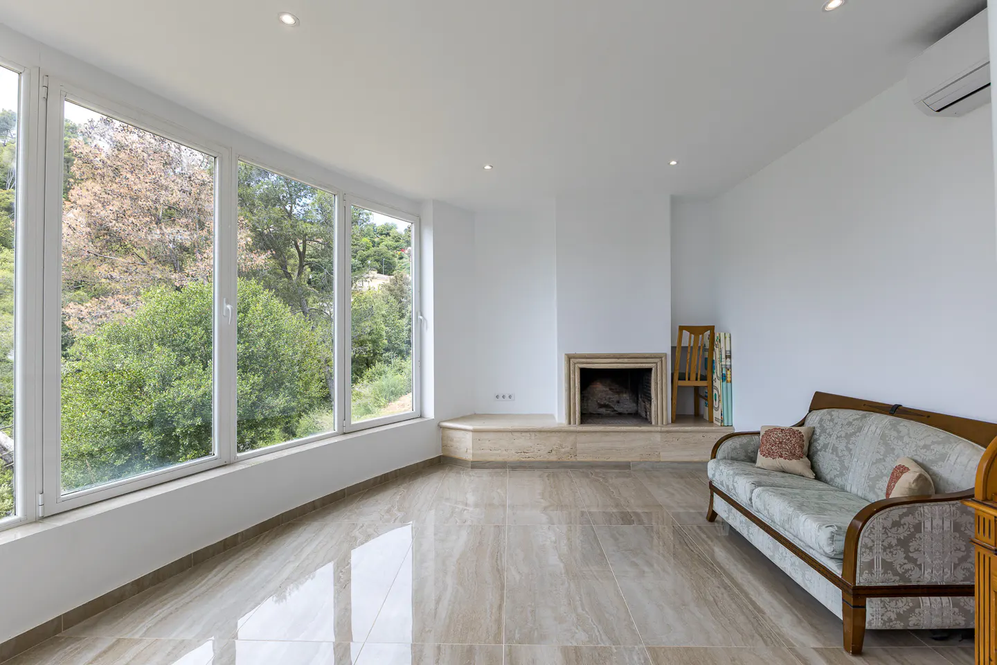 Bright living room with large windows showing trees, a marble fireplace, and a patterned sofa with pillows.
