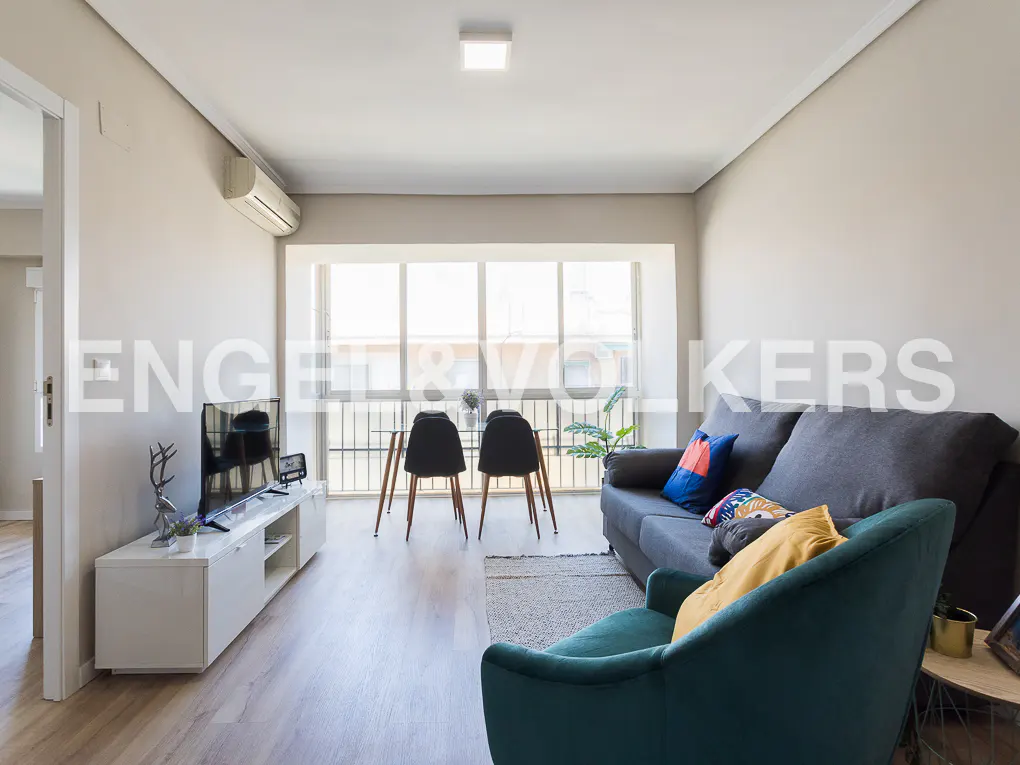 Bright living room with a dark grey sofa, teal armchair, white media console, and a dining area by a large window.