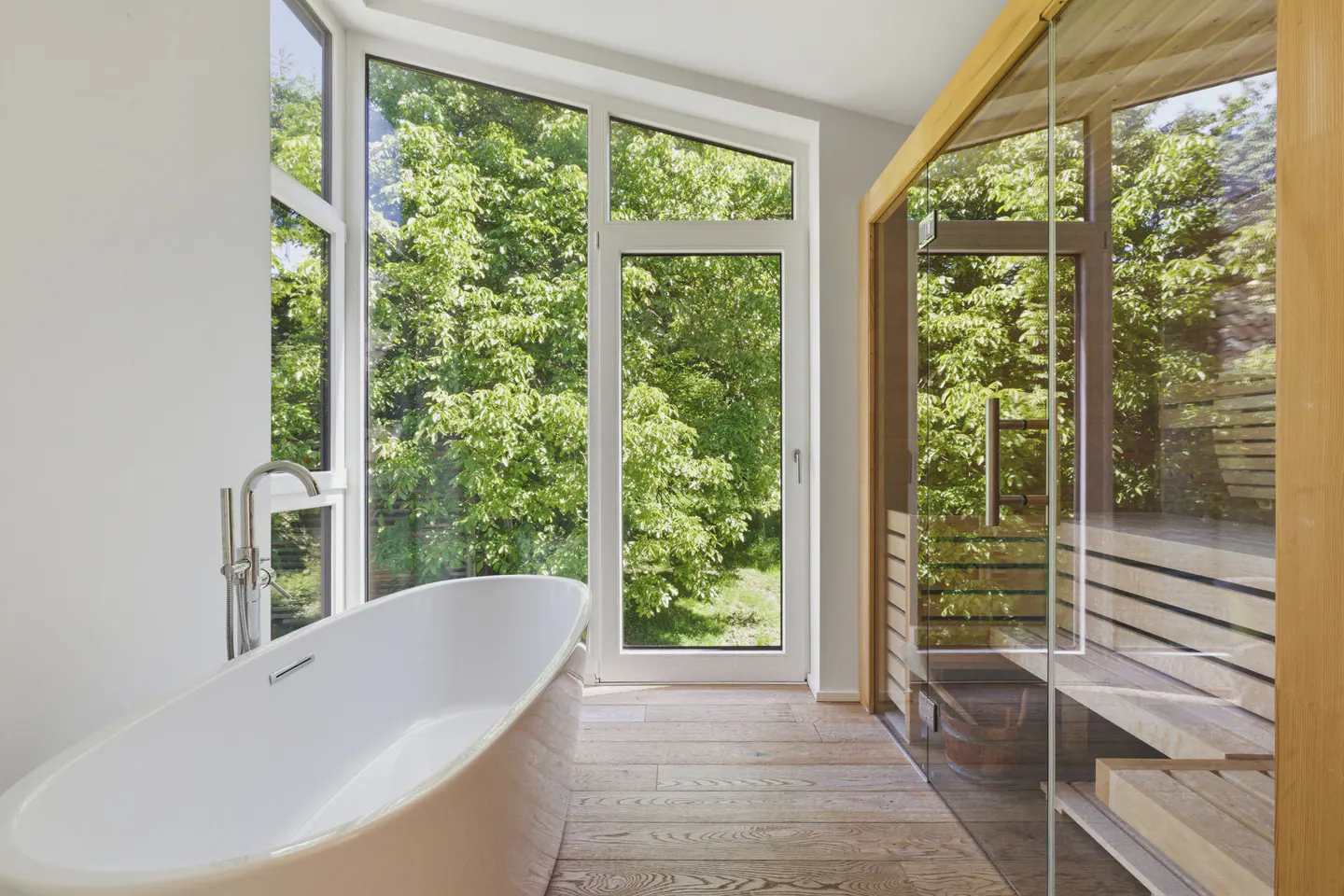 Bright bathroom with a white tub, wood floors, and a glass-enclosed sauna. Large windows overlook lush green trees.