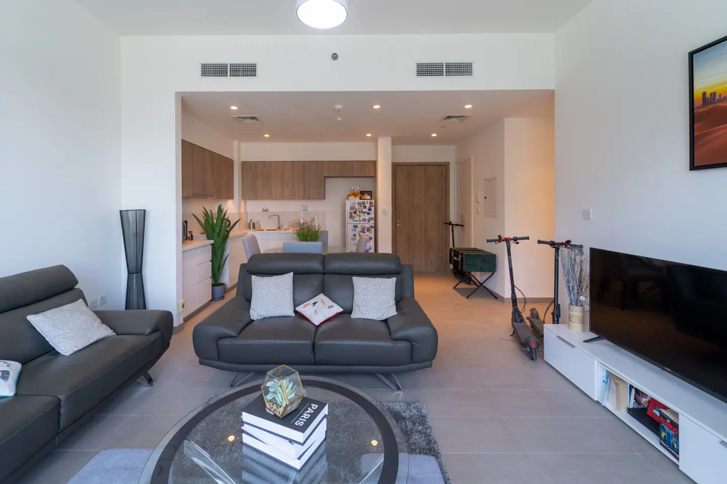 A modern apartment living room with gray sofas, a glass coffee table, and an open kitchen in the background. Two scooters are parked near the TV.