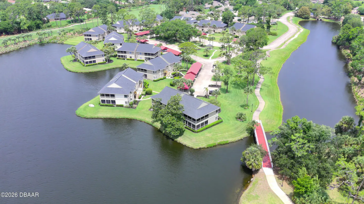 Aerial view of lakeside condos with gray roofs, green lawns, and a red bridge over a narrow waterway.