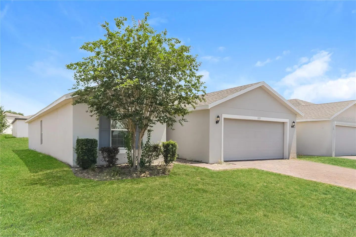 Beige single-story house with a gray garage door, green lawn, and a tree in the front yard under a blue sky.