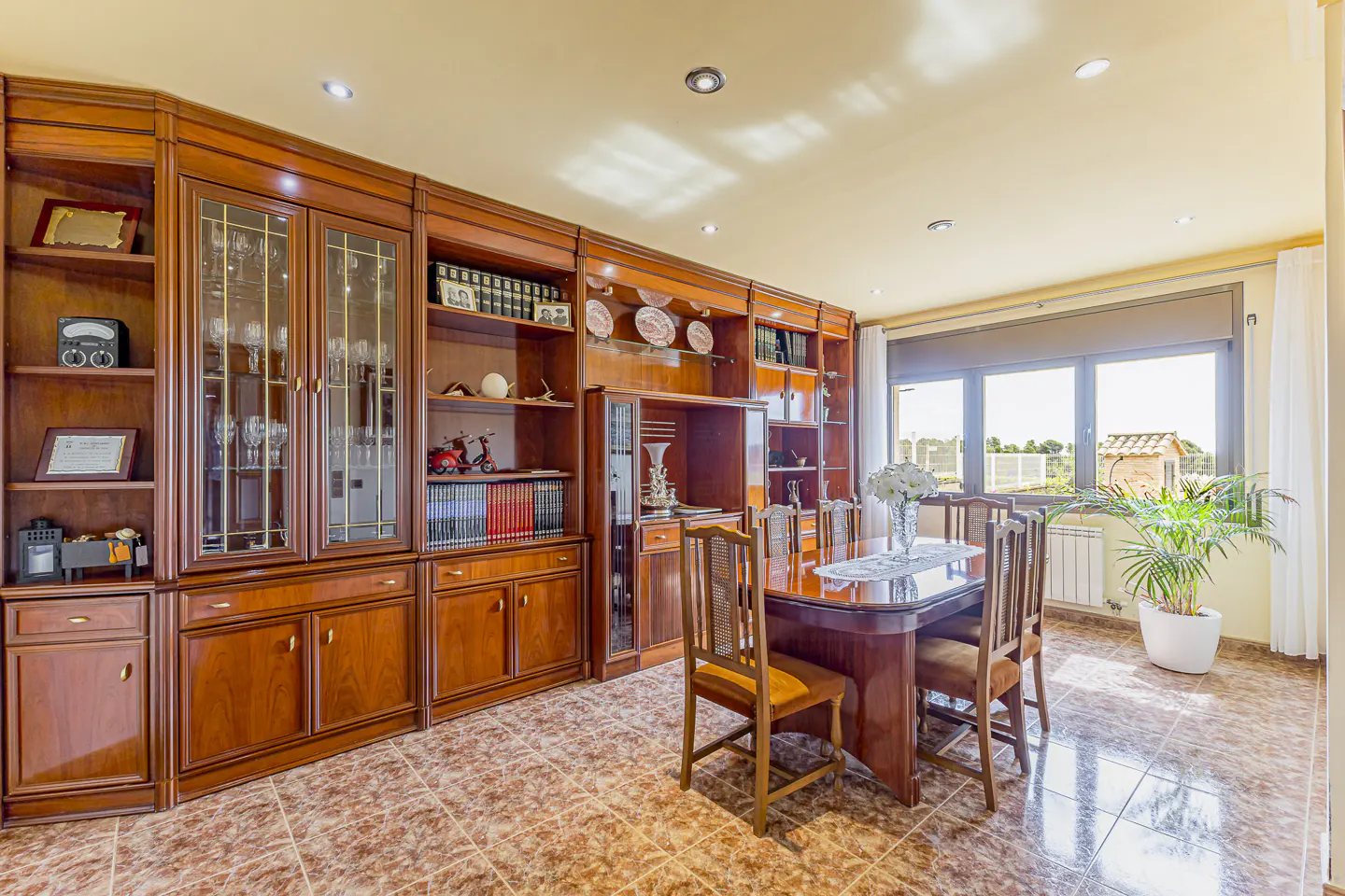Dining room with a large wooden table, chairs, and extensive built-in wooden cabinetry on a tiled floor by a bright window.