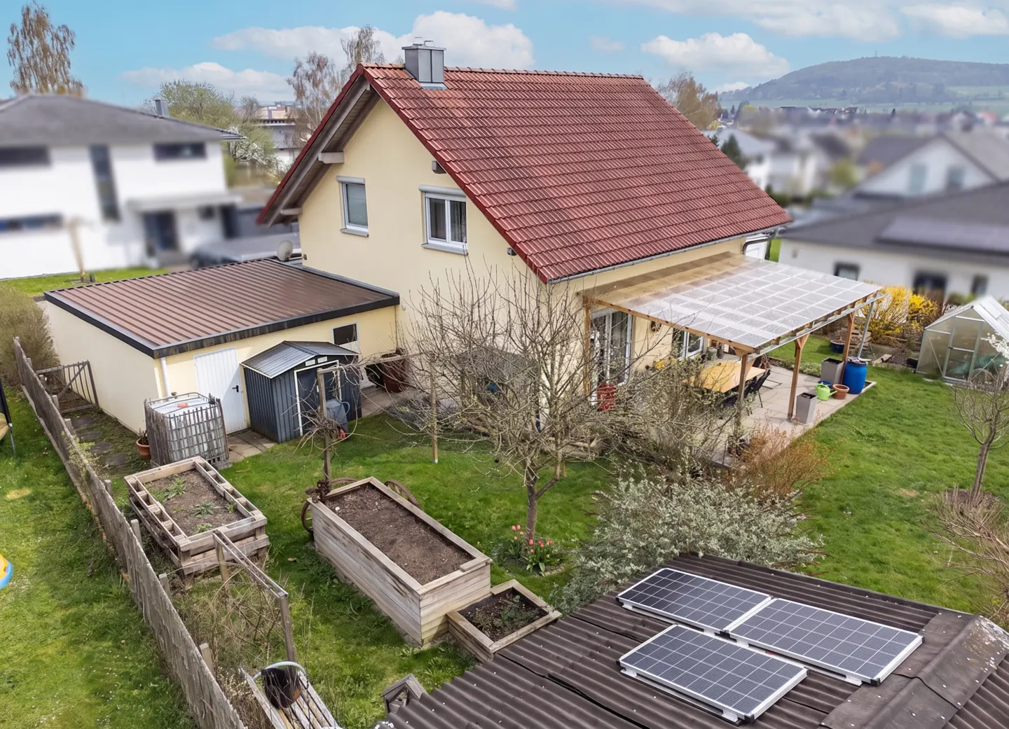Aerial view of a yellow house with a red tile roof, a patio, a green lawn, and solar panels on a shed roof.