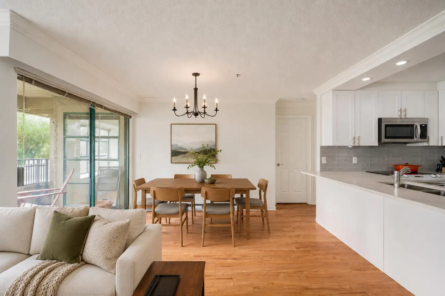 Bright, open-concept living space with a dining table, white kitchen, and a cream-colored sofa. A sliding glass door leads to a balcony.
