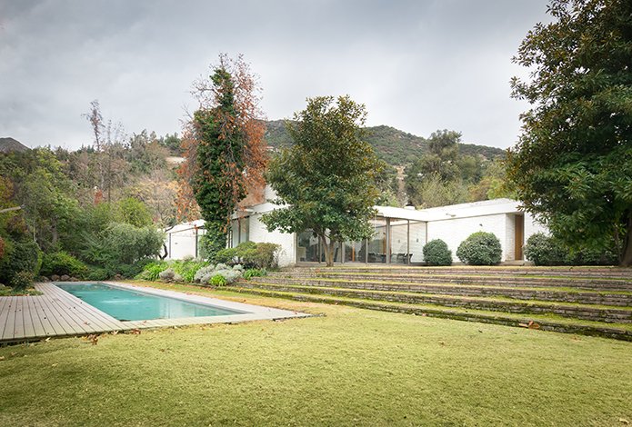 A modern white house with large windows, a pool with a wooden deck, and a green lawn on a cloudy day.