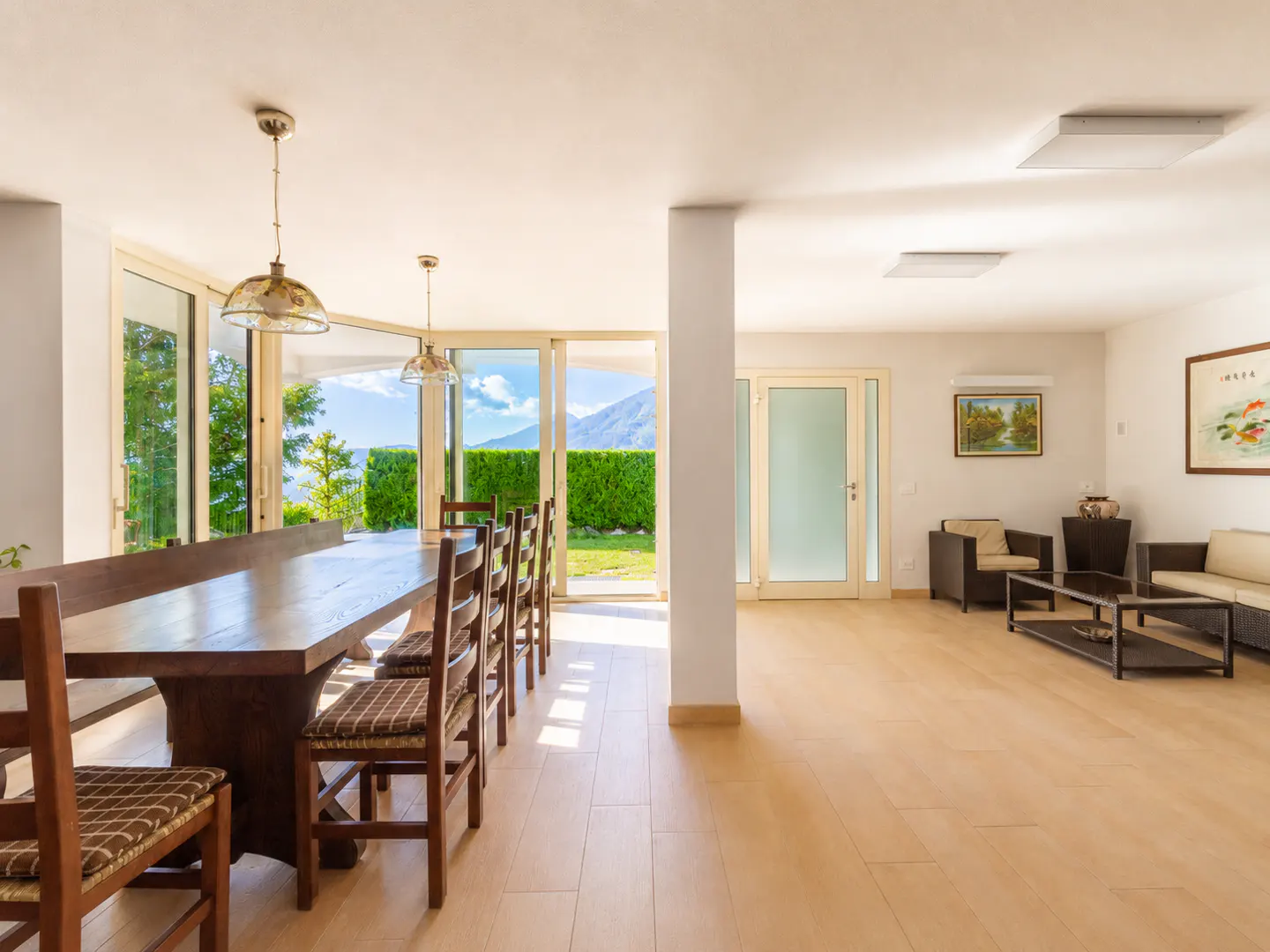 Bright open-plan living area with a wooden dining table, modern seating, and large glass doors overlooking a garden.