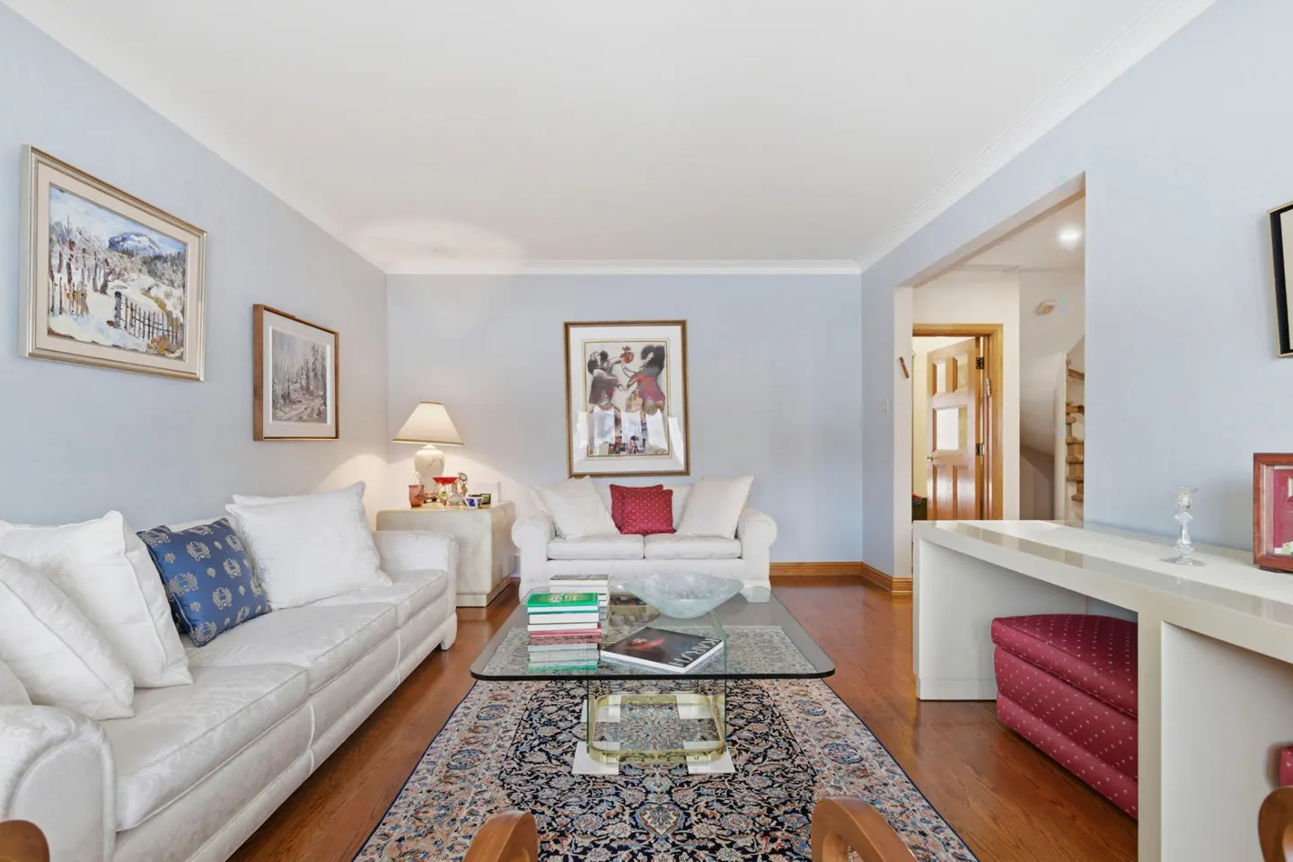 Living room with light blue walls, hardwood floors, and white sofas. A glass coffee table sits on a patterned rug. Artwork adorns the walls.